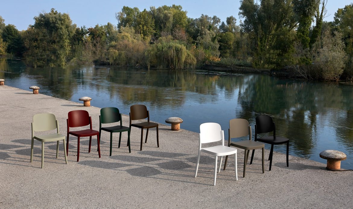 Set of polypropylene chairs from the Piazza collection by Colos in different colours arranged on a concrete pier, with a background of a calm river and lush vegetation.